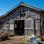 徳島県阿南市にある老朽化した農業用倉庫と古い農機具。青空の下で解体を待つ木造倉庫の様子。