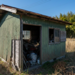 徳島県内の一軒家の庭にある、老朽化して錆びや塗装剥げが見られる古い物置。中には不用品や道具が保管されており、背景には徳島らしい青空と竹林が広がる様子。