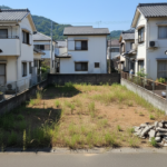 徳島の住宅街にある解体後の一軒家跡地。周囲を住宅に囲まれた狭小地の更地で、地面にはまばらに雑草が生え、隅には解体時のコンクリート瓦礫が積まれている状態。