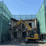 徳島市での住宅解体工事の様子。重機による木造家屋の取り壊しと、近隣への配慮を徹底した高い養生シートの設置。