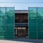 徳島県での解体工事現場。青空の下、2階建ての日本家屋の左右に緑色の養生シートと足場が整然と設置されている様子。