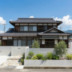 徳島の住宅街にある、築30年の伝統的な木造2階建て住宅の全景。新しくリニューアルされたモダンな外構が特徴で、白い軽量アルミフェンスが、ブロック塀ではなく滑らかなコンクリート擁壁の上に設置されています。擁壁の内側には、オリーブやアガベ、観賞用グラスなどの清潔感のある植栽が美しく配置され、舗装されたコンクリート駐車場と石畳のアプローチが続いています。背景には周囲の家々と緑豊かな山並みが、澄み切った青空と白い雲の下に広がり、清潔感を表現しています。