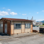 青空の下、徳島の工業地帯に建つ、古く錆びたトタン壁のプレハブ。背景には工場群と山々、クレーンが見える港が広がる。