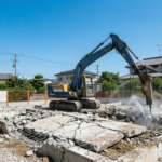 徳島県内の住宅地で青空の下おこなわれているベタ基礎の解体工事風景。油圧ショベルのアタッチメント（ブレーカー）でコンクリート土台を破砕し更地にする様子。