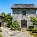 徳島市や阿南市で管理・解体が検討される、伝統的な石造りの蔵（蔵）。青空の下、手入れされた日本庭園の中に立っている。