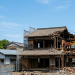 徳島県にある築50年の木造二階建て一軒家の解体工事現場。青空の下、黄色い油圧ショベルが稼働し、木造の骨組みやがれきが見える、リアルで清潔感のある様子。