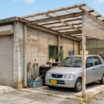 【徳島市・阿南市】築30年ほどの老朽化した車庫とカーポートの下に停まる軽自動車。コンクリートブロックとトタン屋根の古びた構造で、周囲は典型的な徳島の住宅景観。