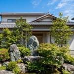 青空の下、庭石と多様な植栽（松、カエデ、ツツジなど）が美しく配置された和風庭園を持つ、瓦屋根の現代的な日本の住宅の外観。