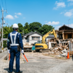 「青空の下、徳島県の住宅街にある解体工事現場で、交通誘導灯を手に持ち、白いヘルメットと紺色の作業服、反射ベストを着用して立つ警備員の後ろ姿。奥では黄色のショベルカーが住宅を解体している。」